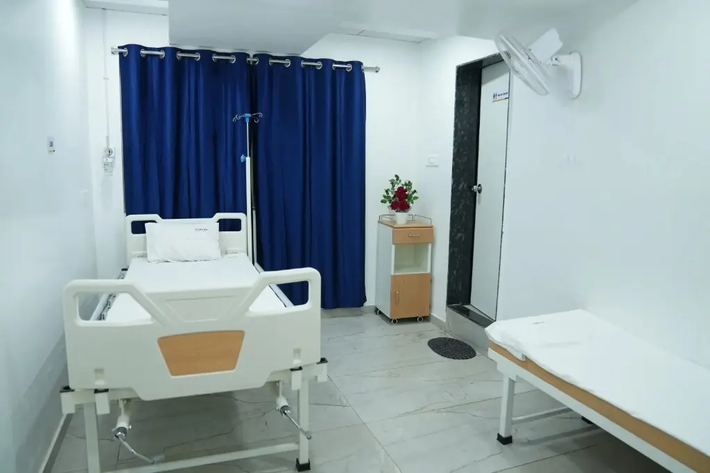 Bright private patient room at Sharvari Hospital with adjustable hospital bed, bedside cabinet, IV stand, and comfortable furnishings.